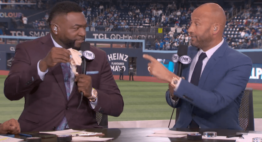 Two men in suits at a broadcasting desk, one holding a small, light-colored cloth, the other pointing and looking at him.